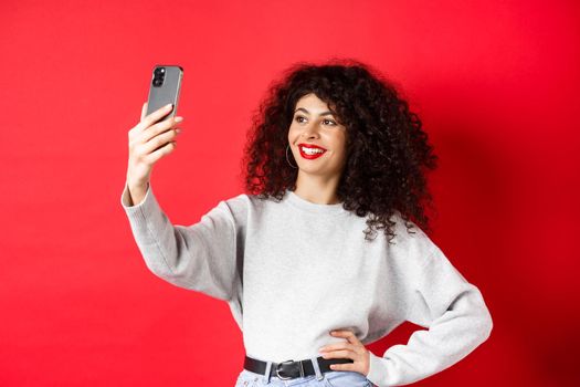 Image of stylish female blogger taking selfie on smartphone, posing for photo on mobile phone, standing at red background.