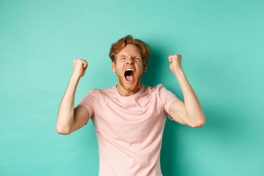 Handsome redhead man watching sports game and cheering, shouting and making fist pumps, looking at screen and watching competition, rooting for team, standing over turquoise background.