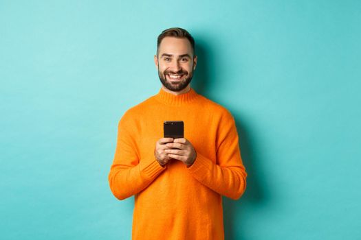 Happy handsome man writing message on mobile phone, holding smartphone and smiling, standing against turquoise background.
