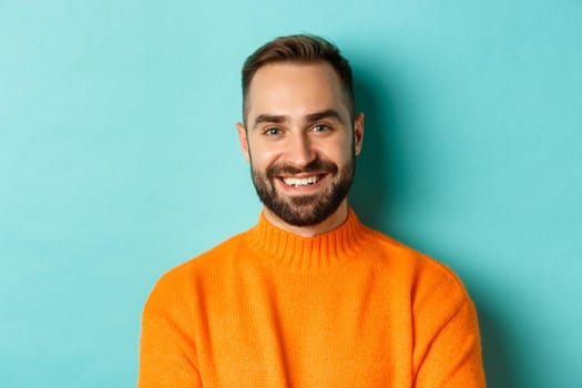 Close-up of handsome caucasian man smiling at camera, looking confident, wearing orange sweater, standing against turquoise background.