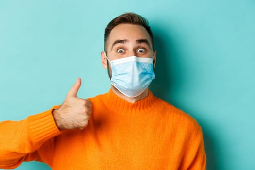 Covid-19, social distancing and quarantine concept. Close-up of cheerful man in medical mask showing thumb-up, light blue background.