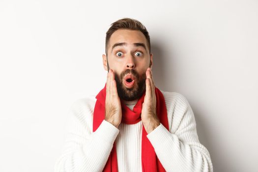 Christmas holidays. Close-up of surprised bearded guy saying wow, holding hands near face, standing against white background.