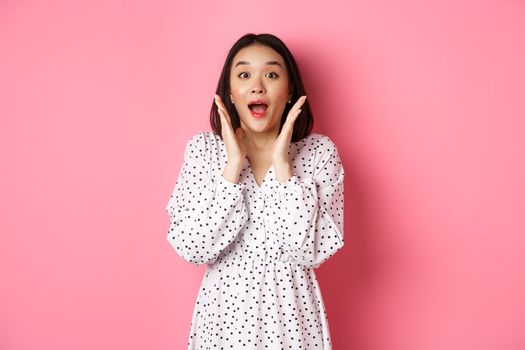 Image of surprised korean girl in dress, female model staring at camera and gasping amazd, standing over pink background.