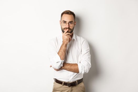 Thoughtful handsome businessman looking at camera, making choice or thinking, standing in glasses and white collar shirt against studio background.