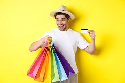 Concept of vacation and finance. Happy man shopper looking at shopping bags satisfied, showing credit card, standing against yellow background.