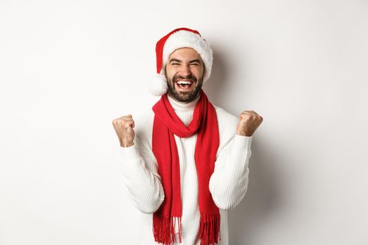 Christmas holidays. Man winner celebrating and triumphing, raising hands up in rejoice, achieve goal, wearing Santa hat and red scarf, white background.