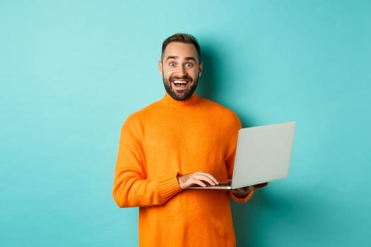 Excited smiling man working on laptop, staring at camera happy, standing in orange sweater against turquoise background.