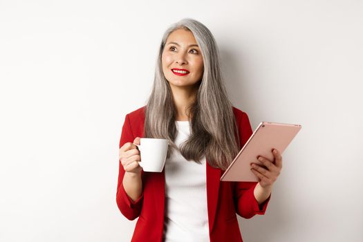 Business people concept. Asian businesswoman drinking coffee and holding digital tablet, smiling thoughtful at upper left corner, white background.