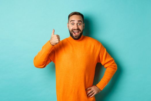 Well done. Handsome bearded man showing thumb up, praising good work, recommend excellent product, like and approve, smiling satisfied, standing over light blue background.