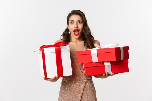 Holidays, celebration concept. Excited and surprised woman holding Christmas gifts and smiling amazed, wearing glamour dress, standing over white background.