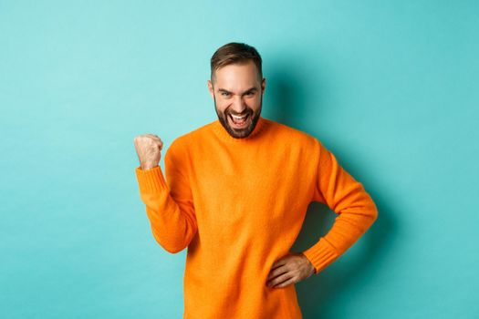 Image of satisfied young man feeling winner, fist pump and saying yes with satisfaction, achieve goal, triumphing while standing over light blue background.