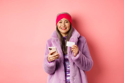 Online shopping and fashion concept. Smiling middle-aged woman in stylish fur coat using mobile phone and plastic credit card, standing happy on pink background.
