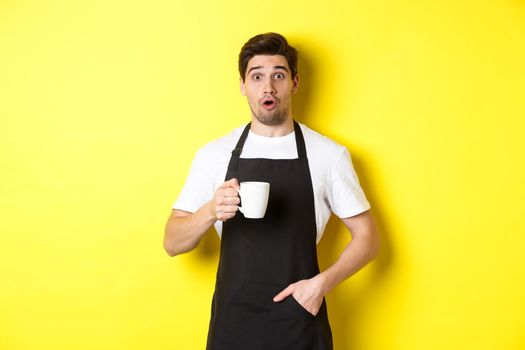 Barista holding coffee mug and looking surprised, standing in black apron cafe uniform against yellow background.