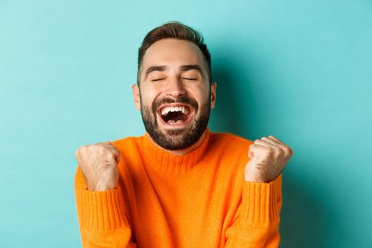 Image of handsome relieved man feeling satisfaction, rejoicing of winning or achievement, making fist pump and saying yes, standing over turquoise background.