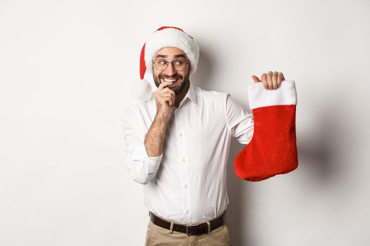 Merry christmas, holidays concept. Adult man looking happy and curious at xmas sock, receive gifts, wearing santa hat, white background.