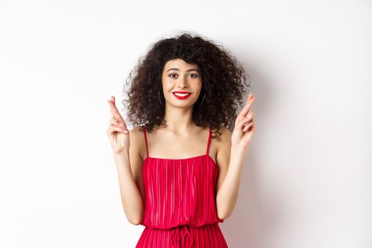 Hopeful beauty girl with curly hair, cross fingers for good luck and smiling, making wish or praying, standing on white background.