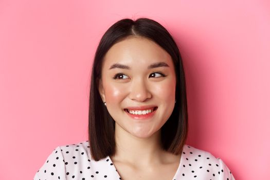 Beauty and lifestyle concept. Headshot of pretty asian woman dreamy gazing left at copy space, smiling happy, standing over pink background.