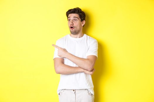 Impressed man in white t-shirt, looking and pointing finger left at promo, check out advertisement, standing against yellow background.