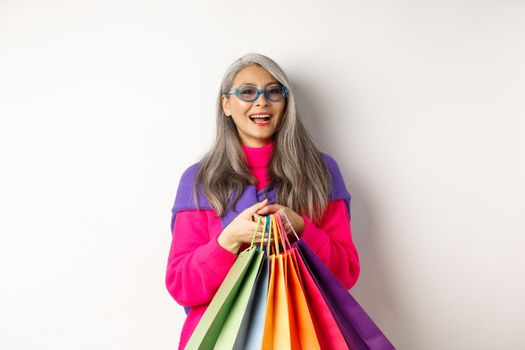 Stylish senior asian woman in sunglasses going shopping on holiday sale, holding paper bags and smiling, standing over white background.