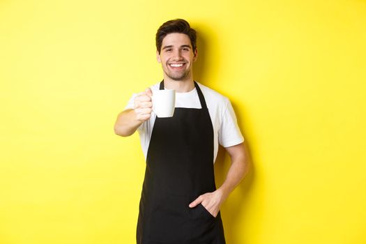 Handsome barista in black apron giving you cup of coffee and smiling, standing over yellow background.