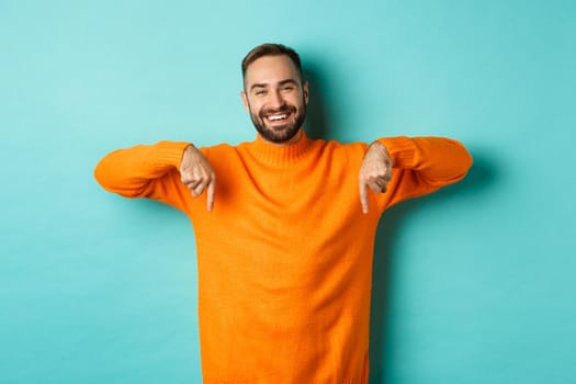 Happy adult man with beard pointing fingers down, smiling cheerful, showing advertisement, standing over turquoise background.