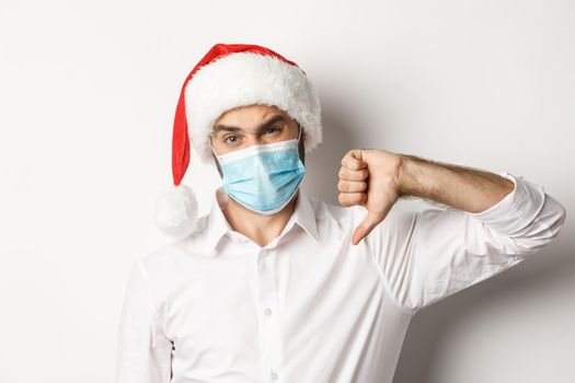 Concept of covid-19, social distancing and winter holidays. Close-up of disappointed man in face mask and christmas hat showing thumb down, standing over white background.