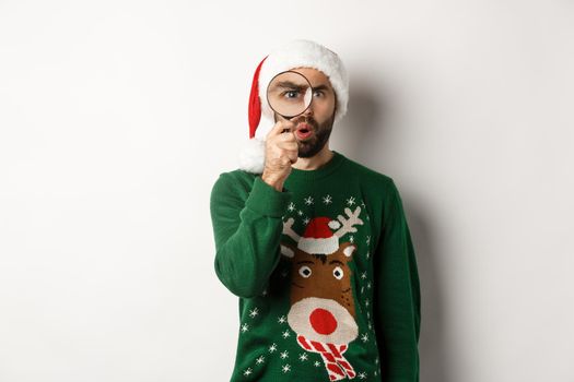 Christmas and holidays concept. Curious guy looking at something through magnifying glass, standing in Santa hat with xmas sweater, standing over white background.