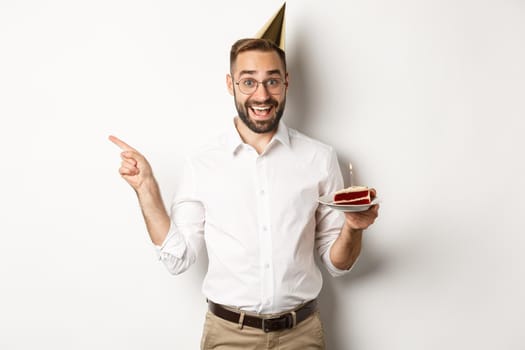 Holidays and celebration. Happy man enjoying birthday party, holding bday cake and pointing finger left at promo, standing over white background.