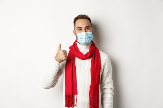 Covid-19, lockdown and quarantine concept. Satisfied smiling man in face mask showing thumb up, approve or like product, standing against white background.