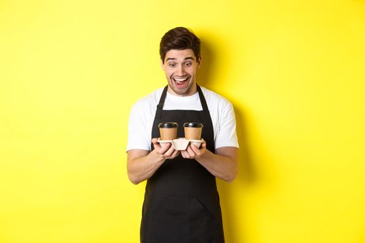 Waiter looking excited at two cups of takeaway coffee, wearing black apron, standing over yellow background.