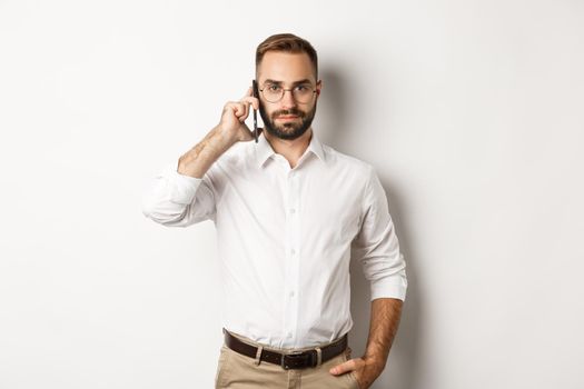 Confident business man talking on phone, looking serious, standing over white background.