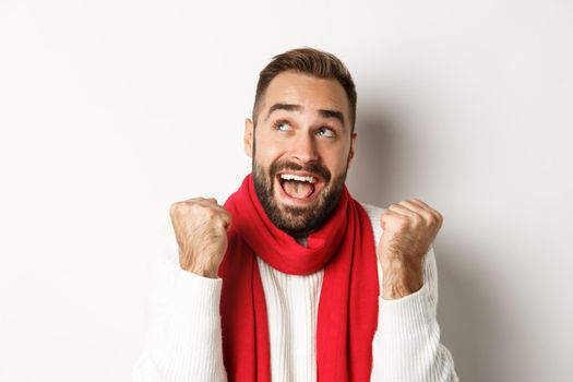 Christmas holidays. Close up of handsome man feeling excited about New Year party, fist pump and looking up amazed, white background.