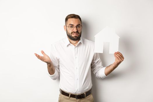 Real estate. Confused man shrugging, showing house paper model and looking indecisive, standing over white background. Copy space