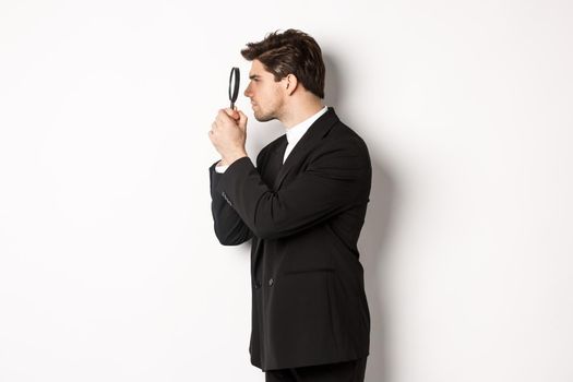 Profile shot of handsome businessman in black suit, looking through magnifying glass and searching for something, standing over white background.