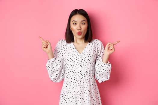 Beautiful asian woman making choice on shopping, pointing fingers sideways and showing variants, smiling at camera, standing over pink background.