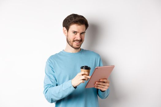 Handsome bearded man smiling at camera, drinking coffee and reading on digital tablet, standing over white background. Copy space