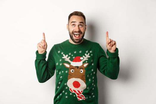 Winter holidays and christmas. Excited bearded man in funny sweater looking, pointing fingers up at copy space, standing over white background.
