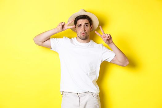 Concept of tourism and vacation. Cool guy taking photo on holidays, posing with peace signs and wearing straw hat, standing against yellow background.