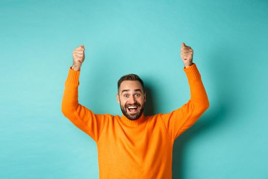 Waist-up shot of happy man raising hands as if holding a sign, showing logo or promo banner, smiling excited, standing in orange sweater against turquoise background.