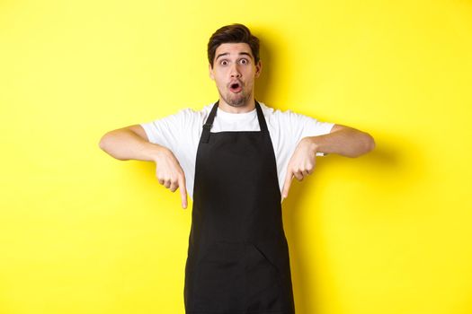 Surprised seller in black apron pointing fingers down, looking amazed at camera, standing over yellow background.