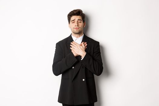 Portrait of dreamy and heartfelt bearded man in suit, close eyes and hold hands on heart, remember something, standing nostalgic over white background.