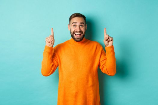 Happy caucasian man showing winter holidays promo offer, pointing fingers up, demonstrate advertisement, standing in sweater against turquoise background.