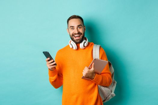 Handsome man student with headphones and backpack, holding digital tablet and smartphone, looking at camera, standing against turquoise background.
