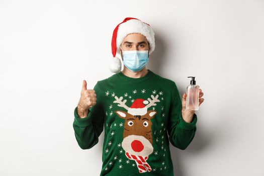 Concept of covid-19 and Christmas holidays. Man recommending hand sanitizer, showing thumb up and antiseptic, wearing medical mask with santa hat, white background.
