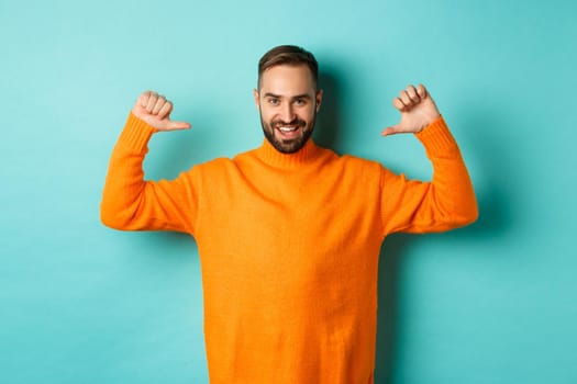 Handsome confident man pointing at himself, looking self-assured, standing in orange sweater against light blue background.