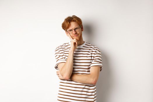 Young man with red messy hair and glasses looking doubtful, thinking and squinting at camera, standing thoughtful over white background.