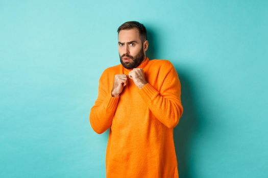 Scared guy jumping startled, looking at something scary, standing in orange sweater, studio background.