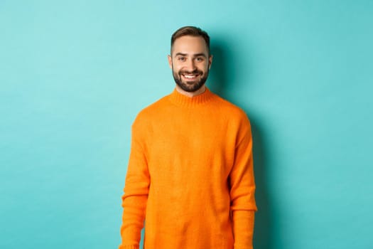 Lifestyle. Handsome man in orange sweater smiling at camera, happy face, guy cheerful expression, standing over turquoise background.