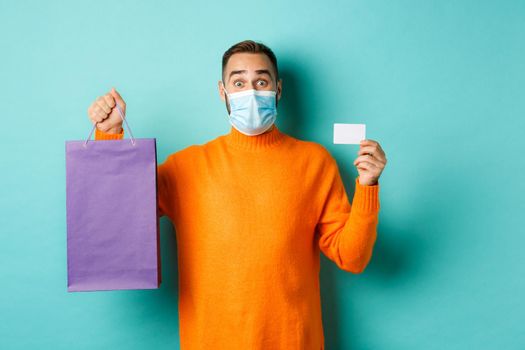 Covid-19, pandemic and lifestyle concept. Happy male customer showing credit card and purple shopping bag, standing over light blue background.