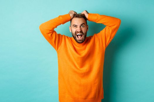 Frustrated man shouting, pulling out hair and screaming angry, losing temper and looking mad, standing over light blue background.